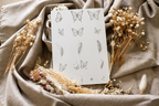Planche de tatouages éphémères papillons et plumes SKNLY présentée sur tissu beige avec fleurs séchées